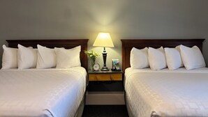Habitación de hotel con dos camas, cada una con ropa de cama y almohadas blancas. Hay una mesita de noche entre las camas con una lámpara, un reloj y un pequeño jarrón con flores.