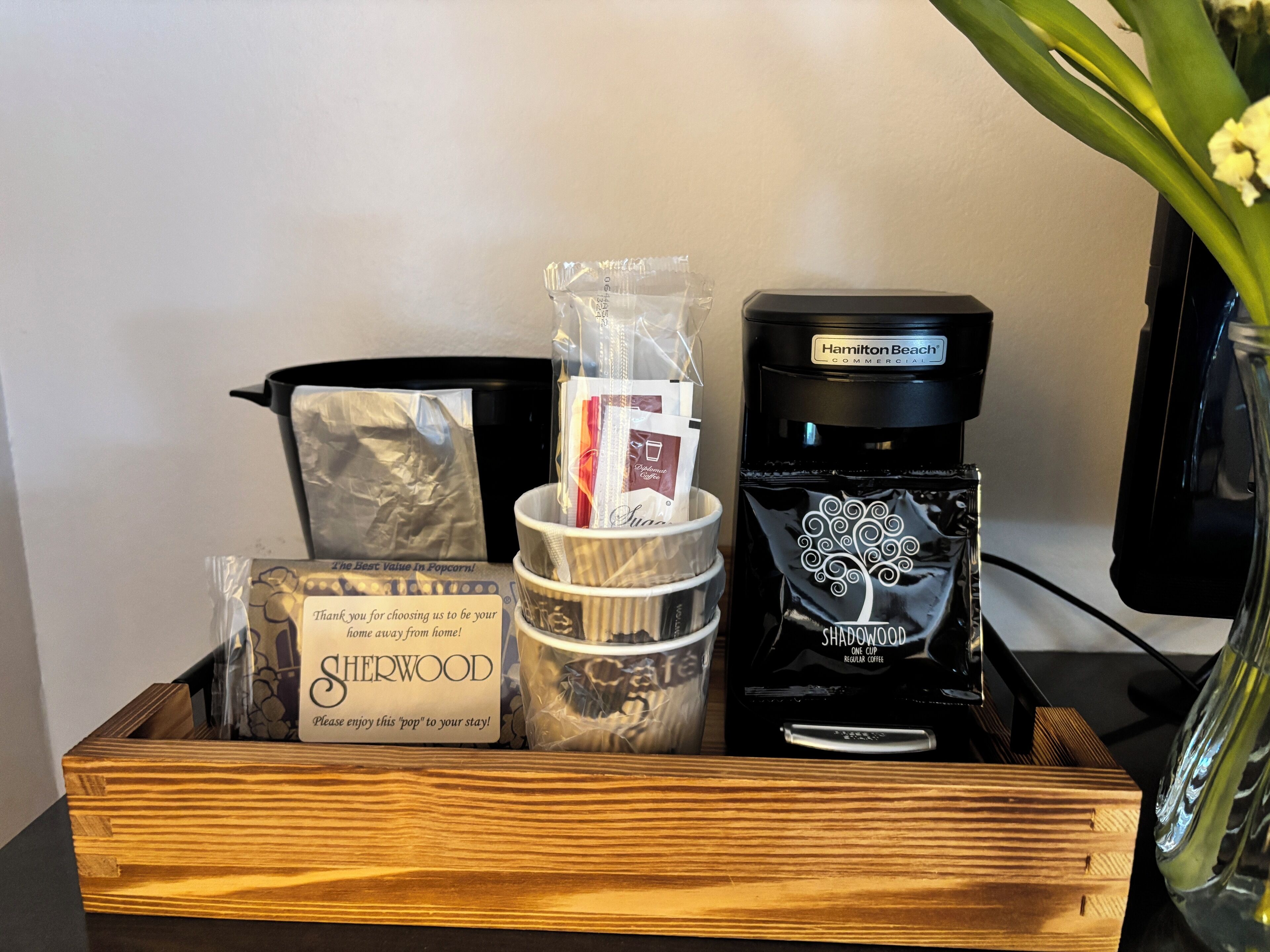A wooden tray with a coffee maker, a bag of Sherwood popcorn, a container of coffee pods, and a glass vase with flowers.