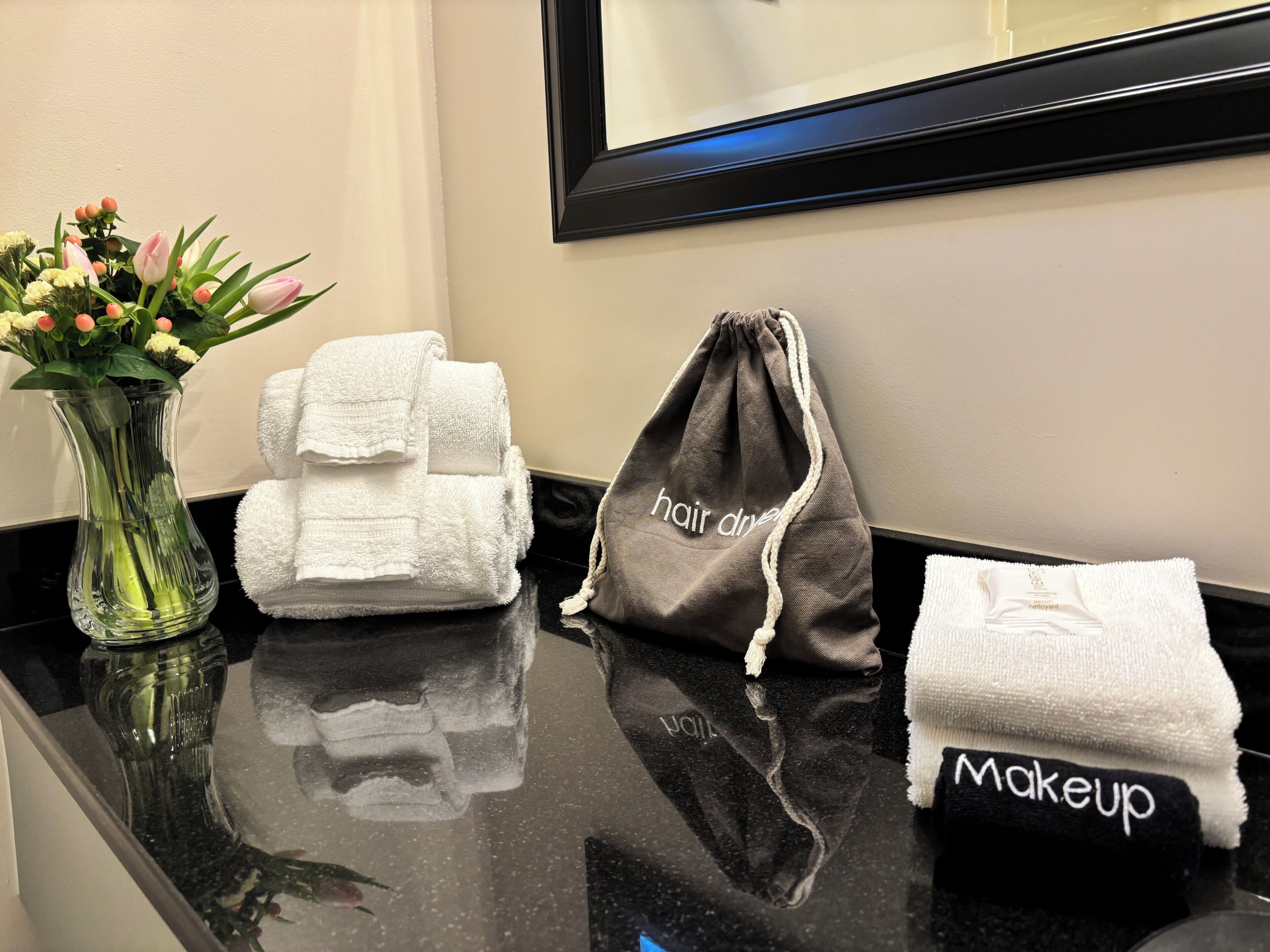 Tocador de baño con un florero, toallas dobladas, una bolsa para secador de pelo y una toalla para maquillaje.