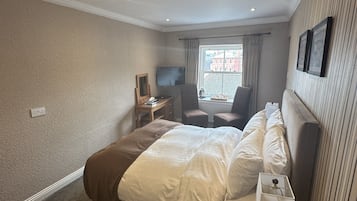 A hotel room with a large bed, a desk with a lamp, a chair, a television, and a window with curtains.