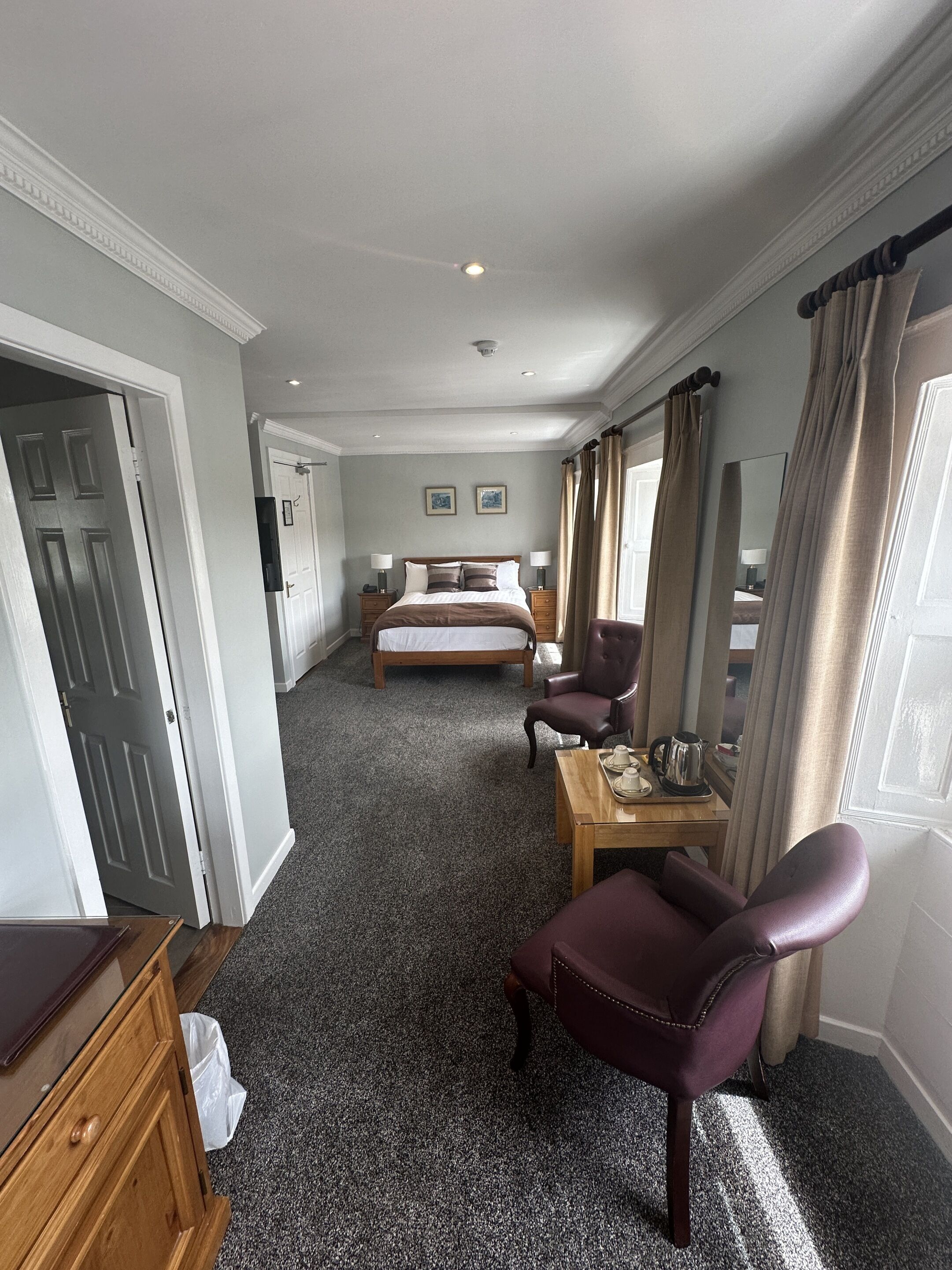 Une chambre d’hôtel avec un grand lit, un bureau avec une chaise, une commode en bois et une porte donnant sur une autre pièce.
