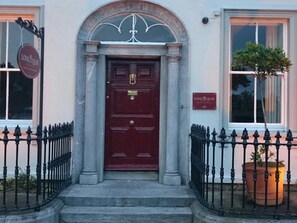 A building with a red door, a sign above the door, and a potted plant to the right. - Long Quay House (Kinsale)