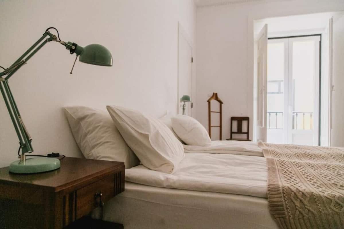 A bedroom with a bed, a nightstand with a lamp, a wooden ladder, and a view of the outside.