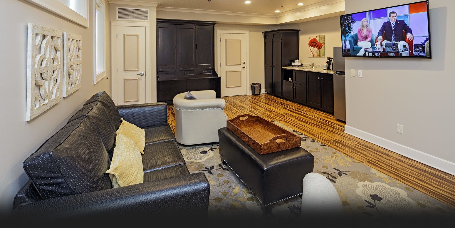 A modern living room with a dark leather sofa, a white ottoman, and a wooden coffee table. There is a kitchen area with dark cabinets and a television mounted on the wall.