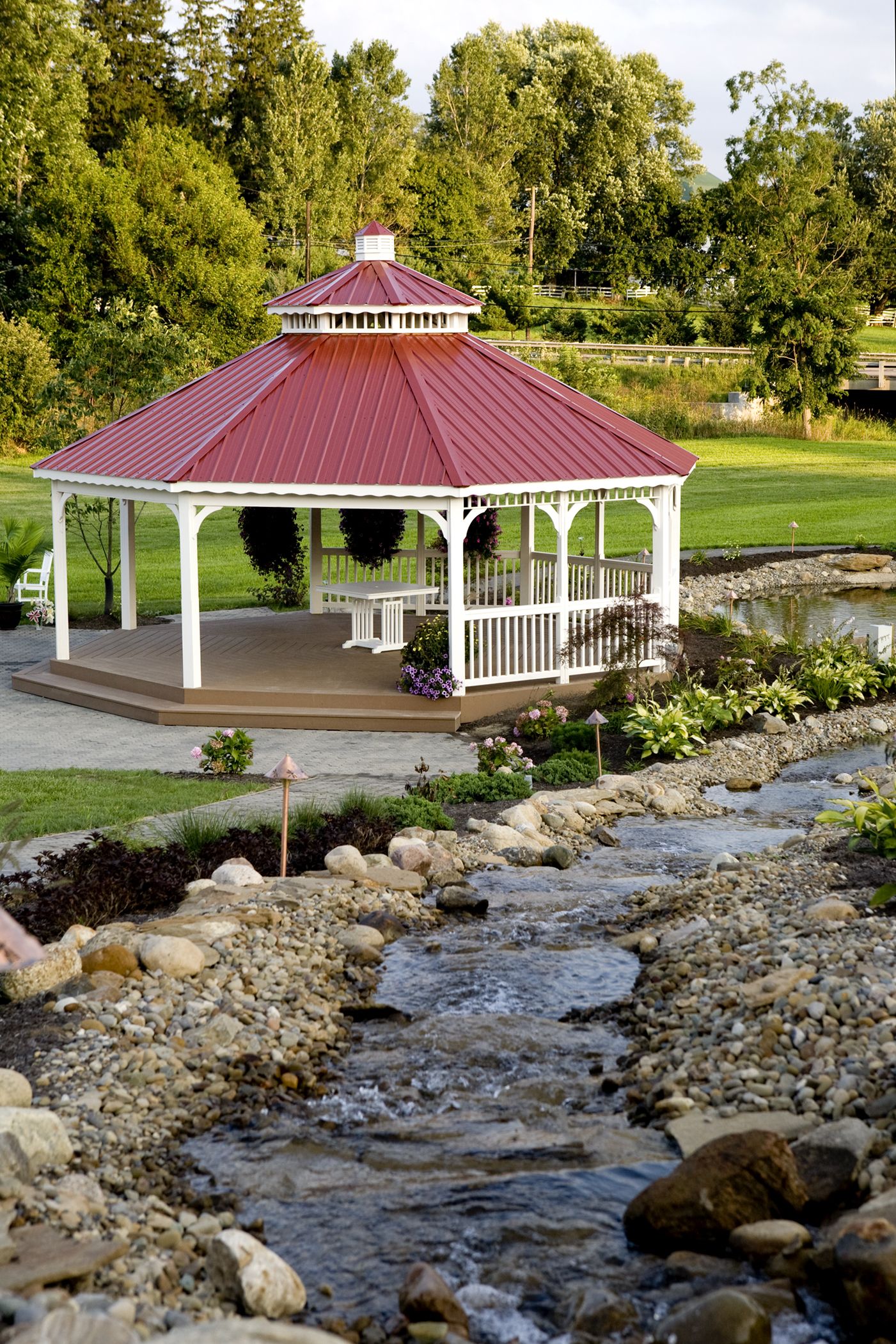 A gazebo with a red roof and white railings, surrounded by a garden with a small pond and pebbles.