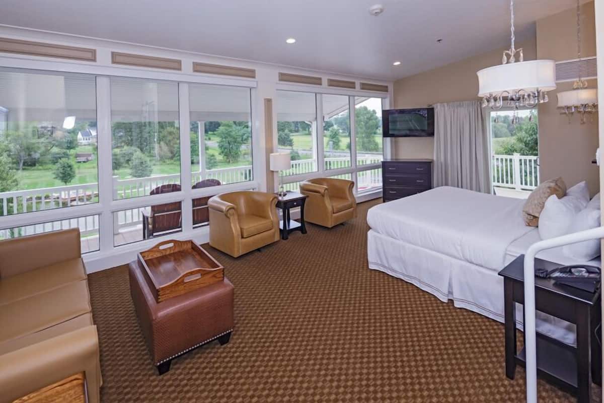 A hotel room with a large bed, two armchairs, a small table, a TV, and a balcony with a view of the greenery.