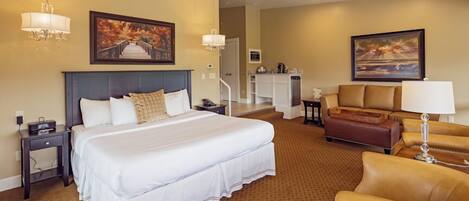 A hotel room with a large bed, a sofa, a desk, and a chair. There is a painting on the wall and a lamp on the desk.