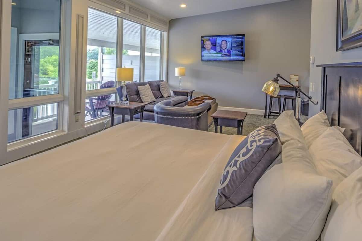 A hotel room with a large bed, a seating area with a sofa and chairs, a television mounted on the wall, and a window providing a view of the outdoors.