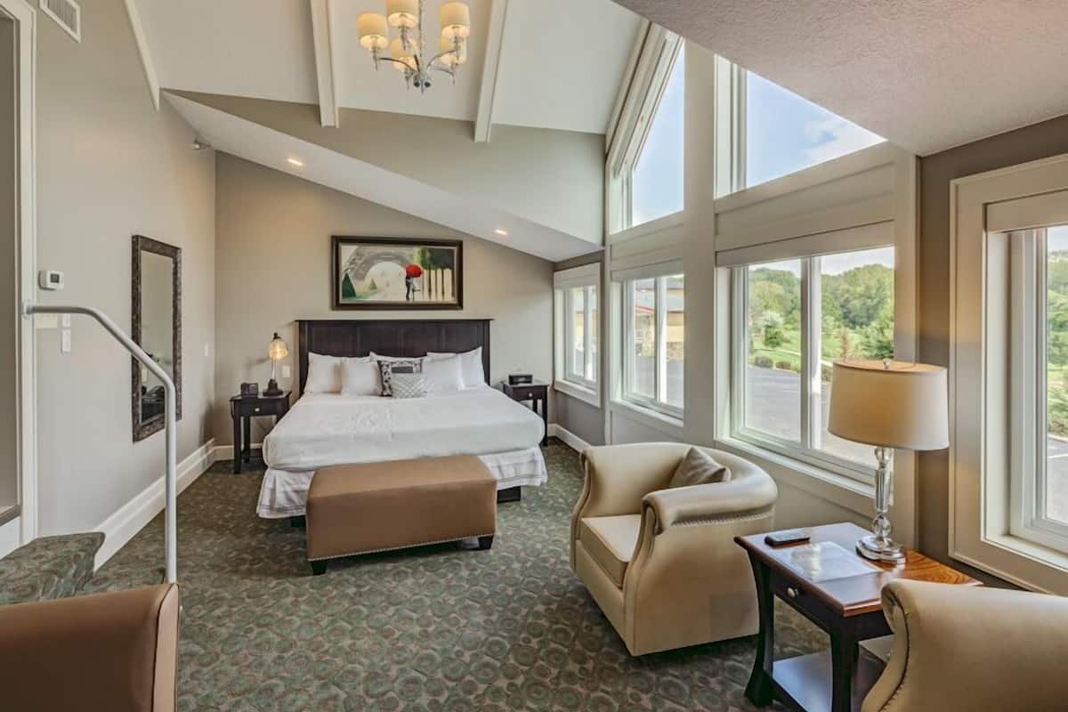 A spacious hotel room with a large bed, a sitting area with a chair and table, and a view of the outdoors through large windows.