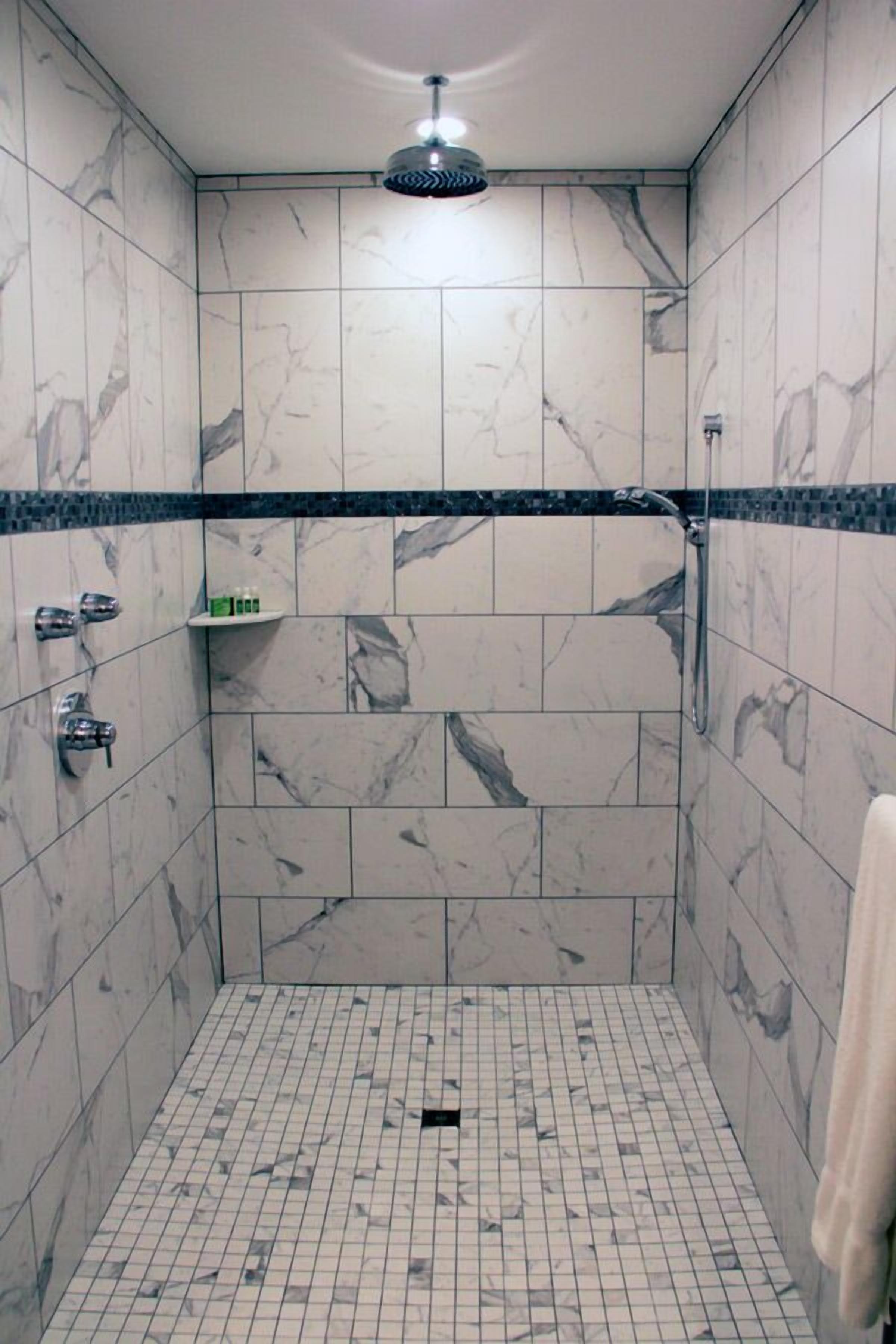 A small, tiled shower with a handheld showerhead, a towel rack, and a shelf with toiletries.