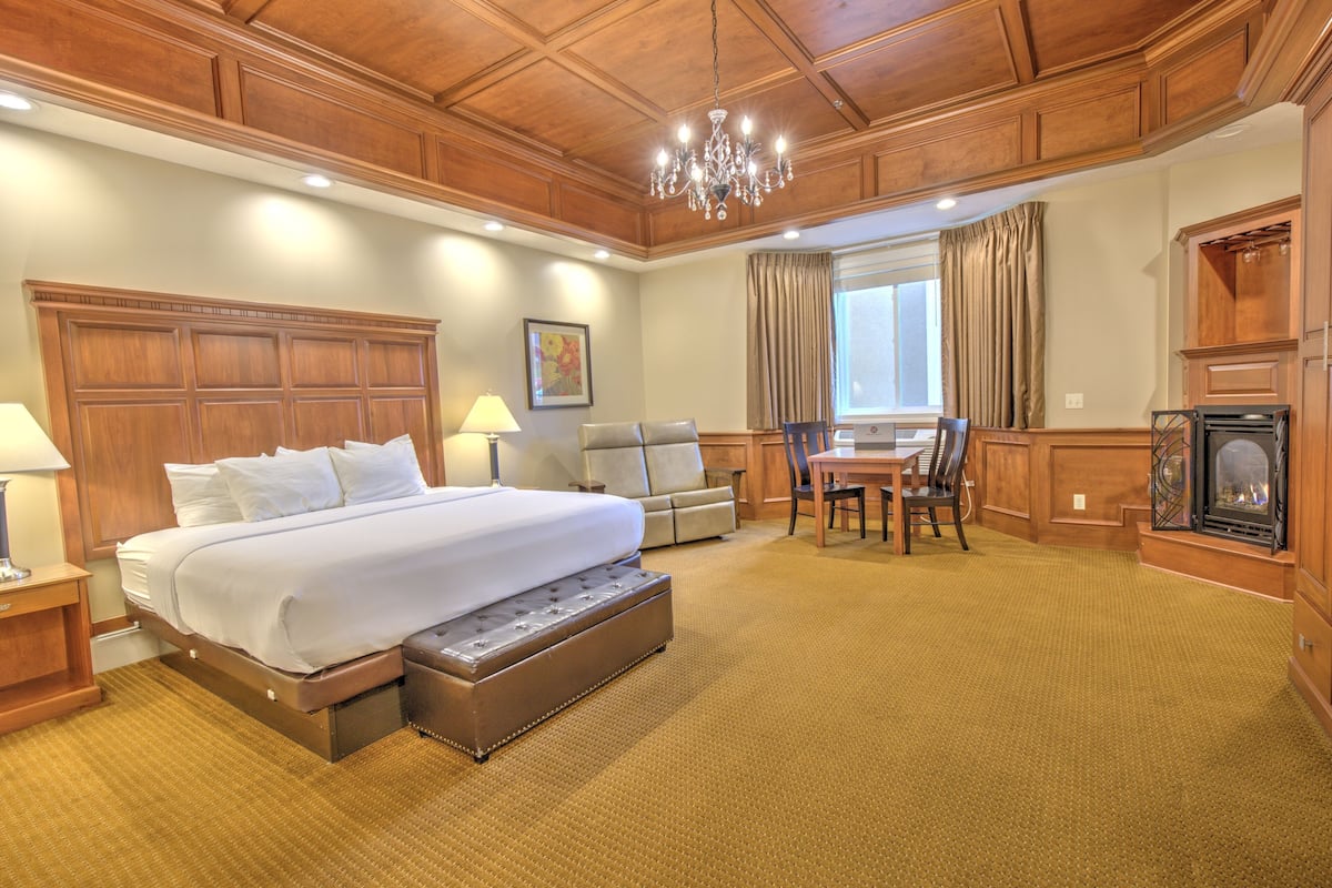A hotel room with a large bed, a desk with chairs, a fireplace, and a chandelier.