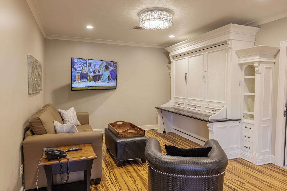 A modern living room with a sofa, ottoman, and a small table with a phone. A TV is mounted on the wall displaying a news program.