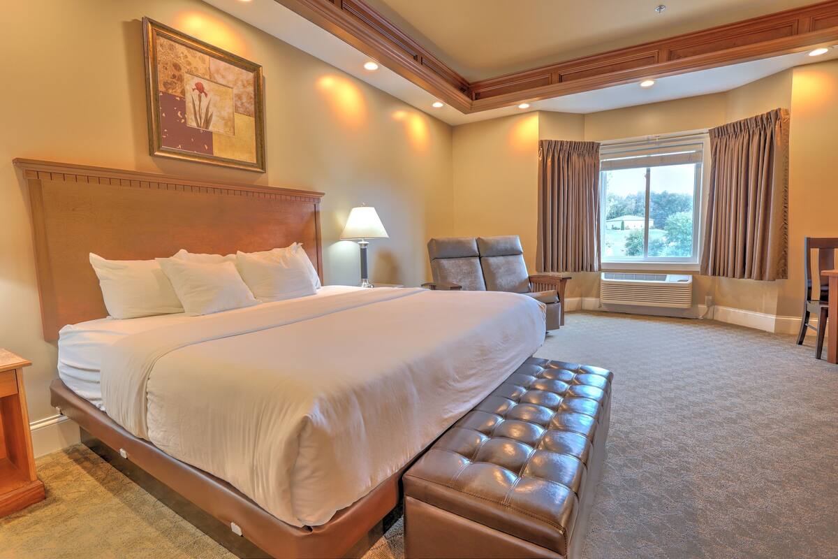 A hotel room with a large bed, a sitting area with a chair, a lamp, and a view of the outdoors through a window.
