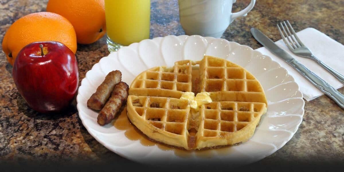A plate with a waffle, sausage, and an apple on a kitchen counter.