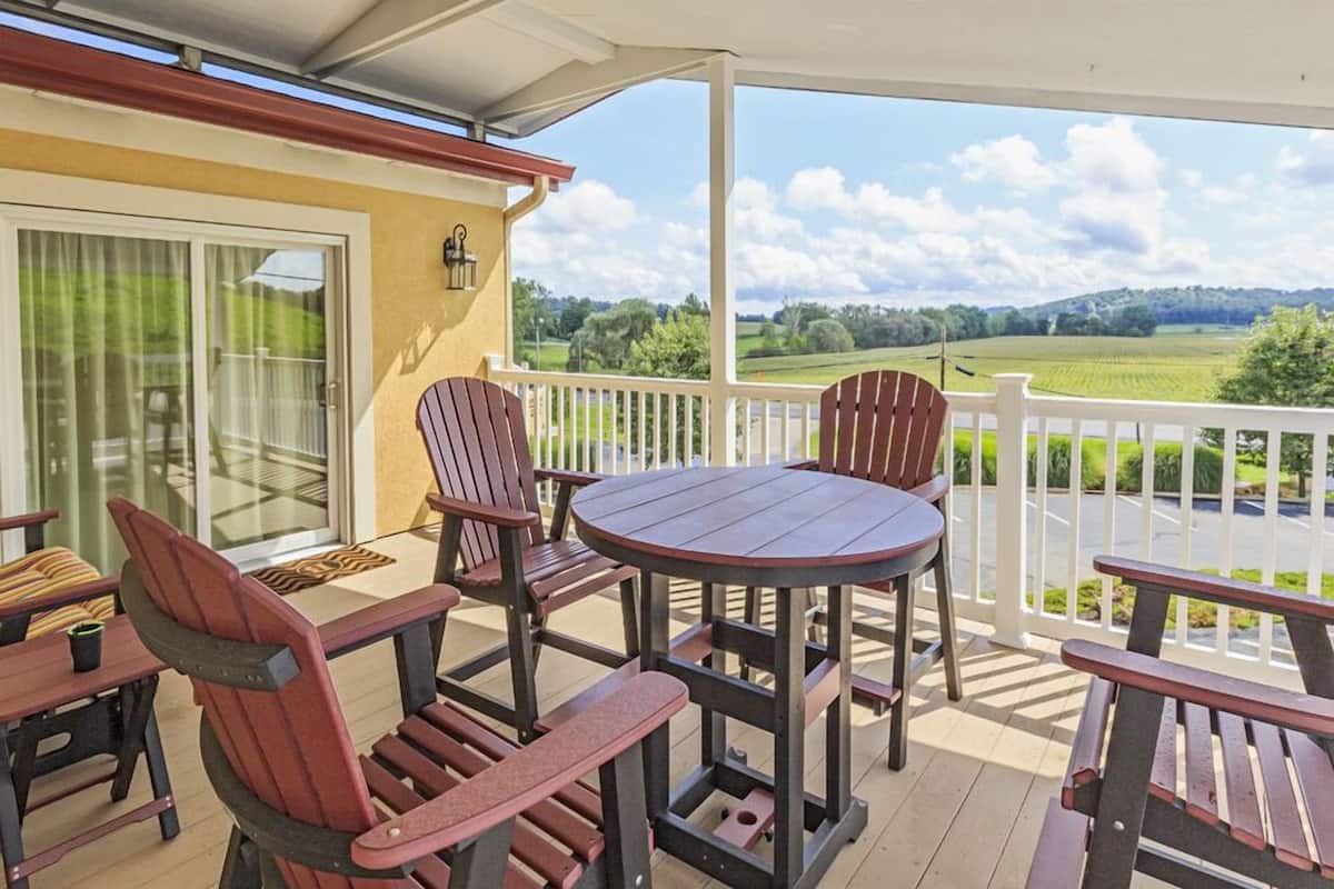 A covered patio with a round table and Adirondack chairs, overlooking a green landscape.