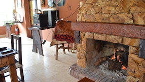 Una chimenea de piedra con fuego, una mesa de madera y una silla en una habitación con piso de baldosas y una pared decorada con diversos objetos.