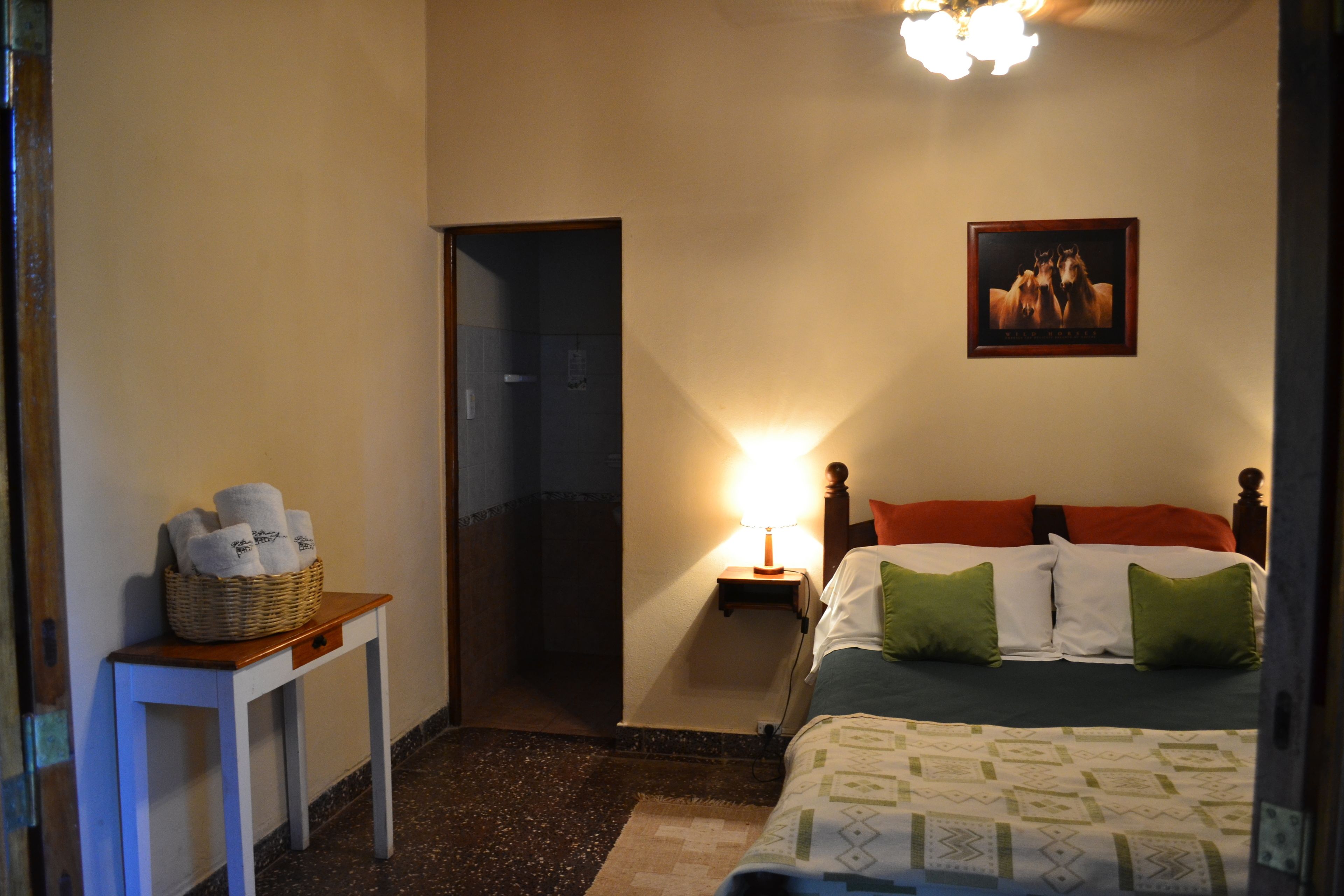 Habitación de hotel con cama, mesita de noche con lámpara, una pequeña mesa con una cesta de toallas y una puerta que da a otra habitación.