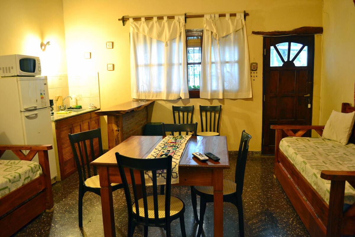 A compact room with a wooden table, chairs, a bed, a microwave, and a small kitchenette.