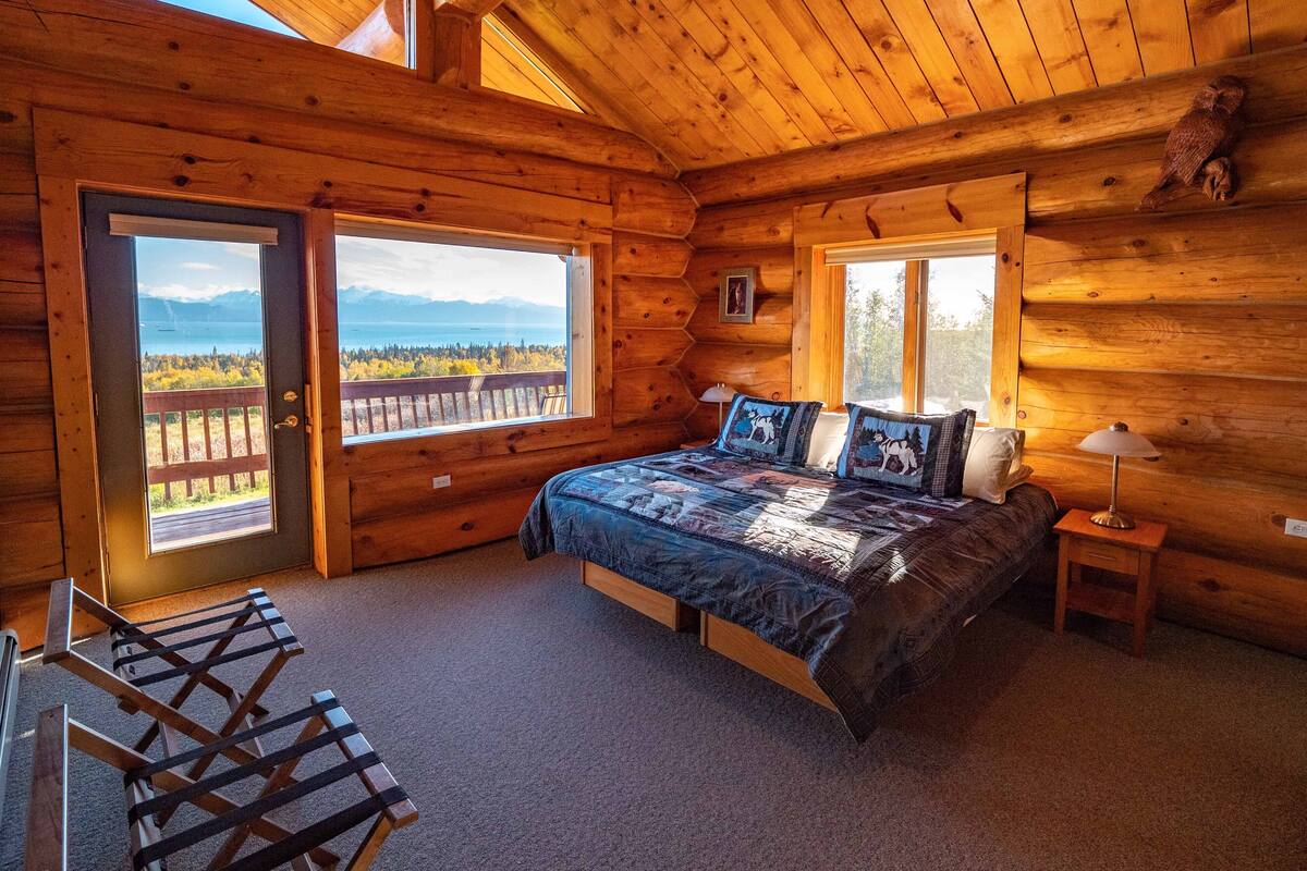 A wooden cabin bedroom with a bed, a nightstand, a lamp, and a view of a lake and mountains through a large window.