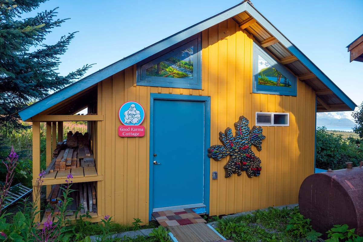 A small wooden cottage with a blue door, a sign reading "Good Karma Cottage," and a decorative leaf pattern on the wall.