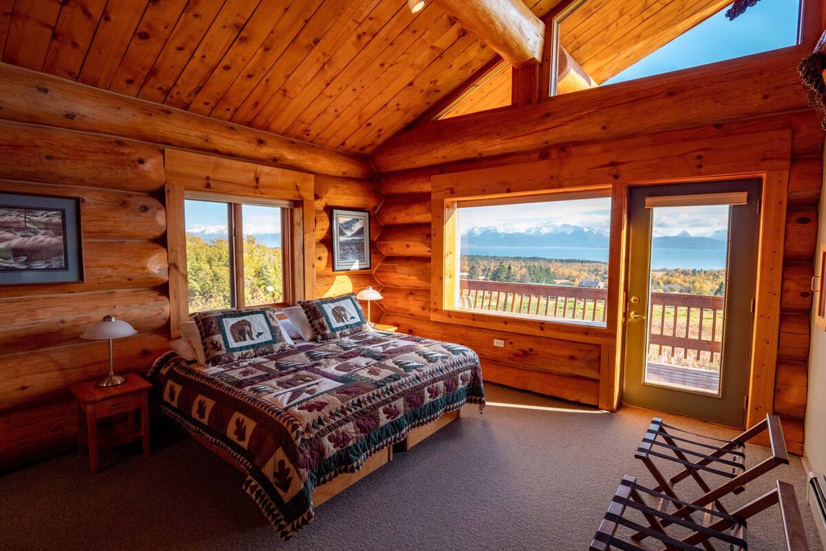 A log cabin bedroom with a bed, a nightstand, a lamp, and a view of mountains and a lake through large windows.