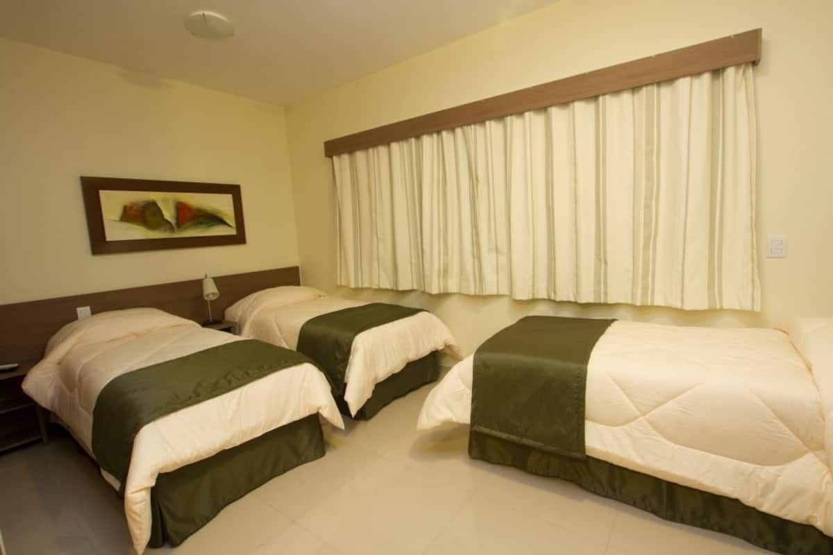 Quarto de hotel com duas camas, mesa de cabeceira, abajur e janela com cortinas.