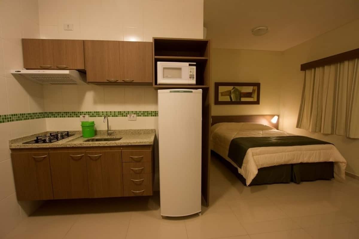 Quarto de hotel compacto com cozinha compacta, incluindo fogão, pia e micro-ondas. Há uma cama com cabeceira e uma luminária de cabeceira.