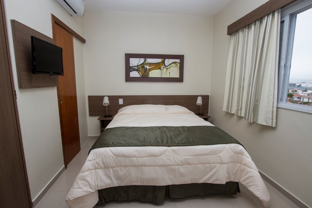 Een hotelkamer met een bed, een flatscreen televisie aan de muur, een schilderij boven het bed, en een raam met gordijnen.