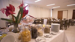 A buffet setup with various food items and condiments in jars, a vase with red flowers, and neatly arranged dining tables and chairs. - My Flat Hotel (Mogi das Cruzes)