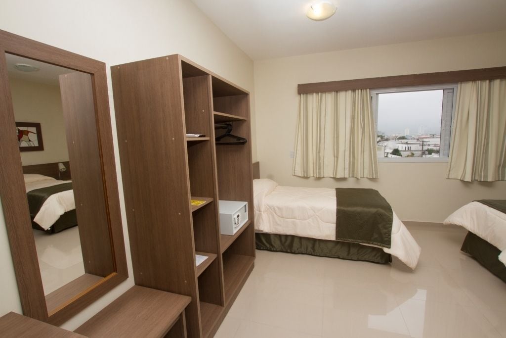 A hotel room with a large mirror, wooden wardrobe, two beds, a window with curtains, and a view of the city.