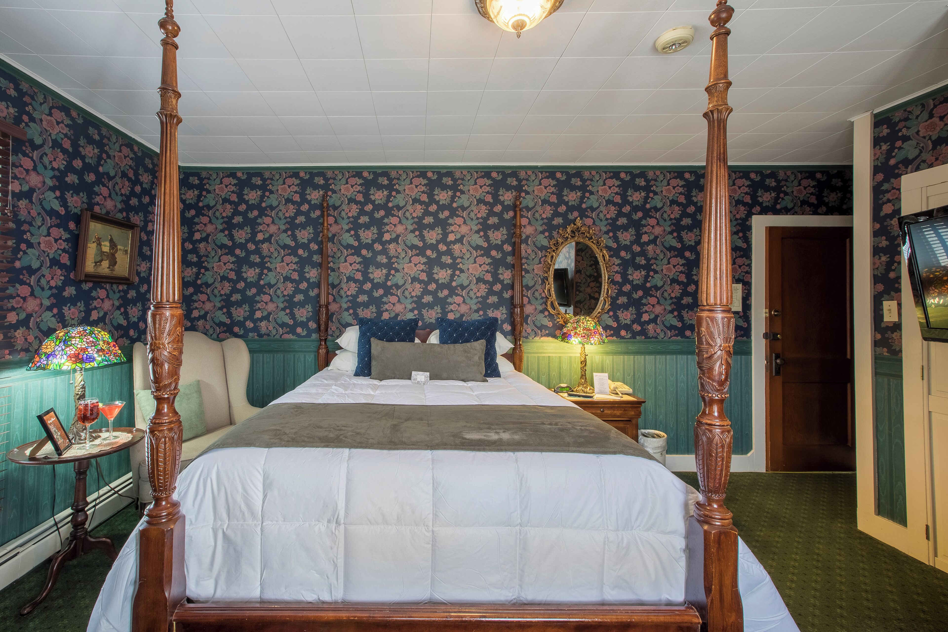 A four-poster bed with a floral wallpaper background, a mirror, and a nightstand with a lamp.