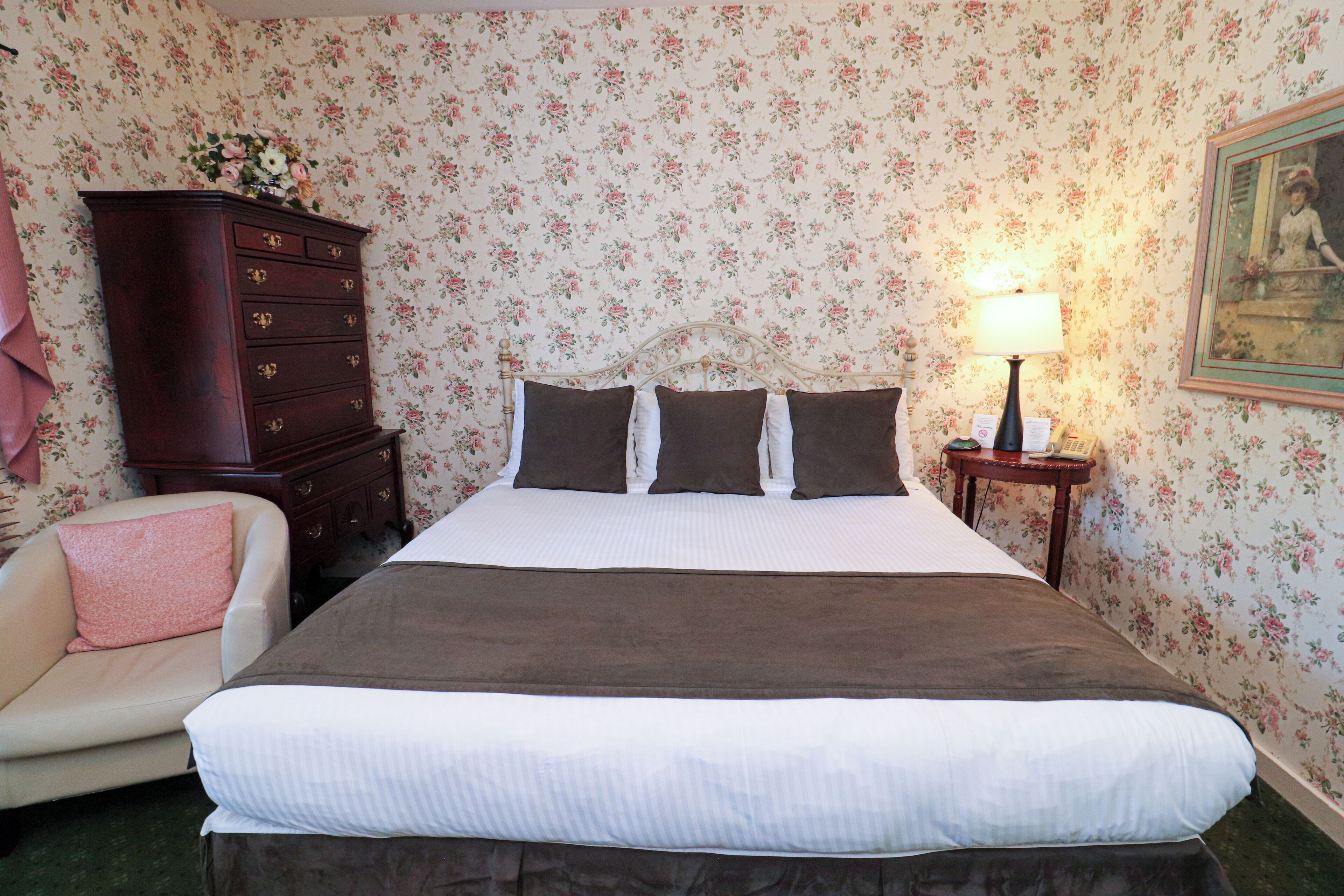 A neatly made bed with a brown bedspread and white linens, a wooden dresser with drawers, a small table with a lamp, and a floral wallpapered wall.