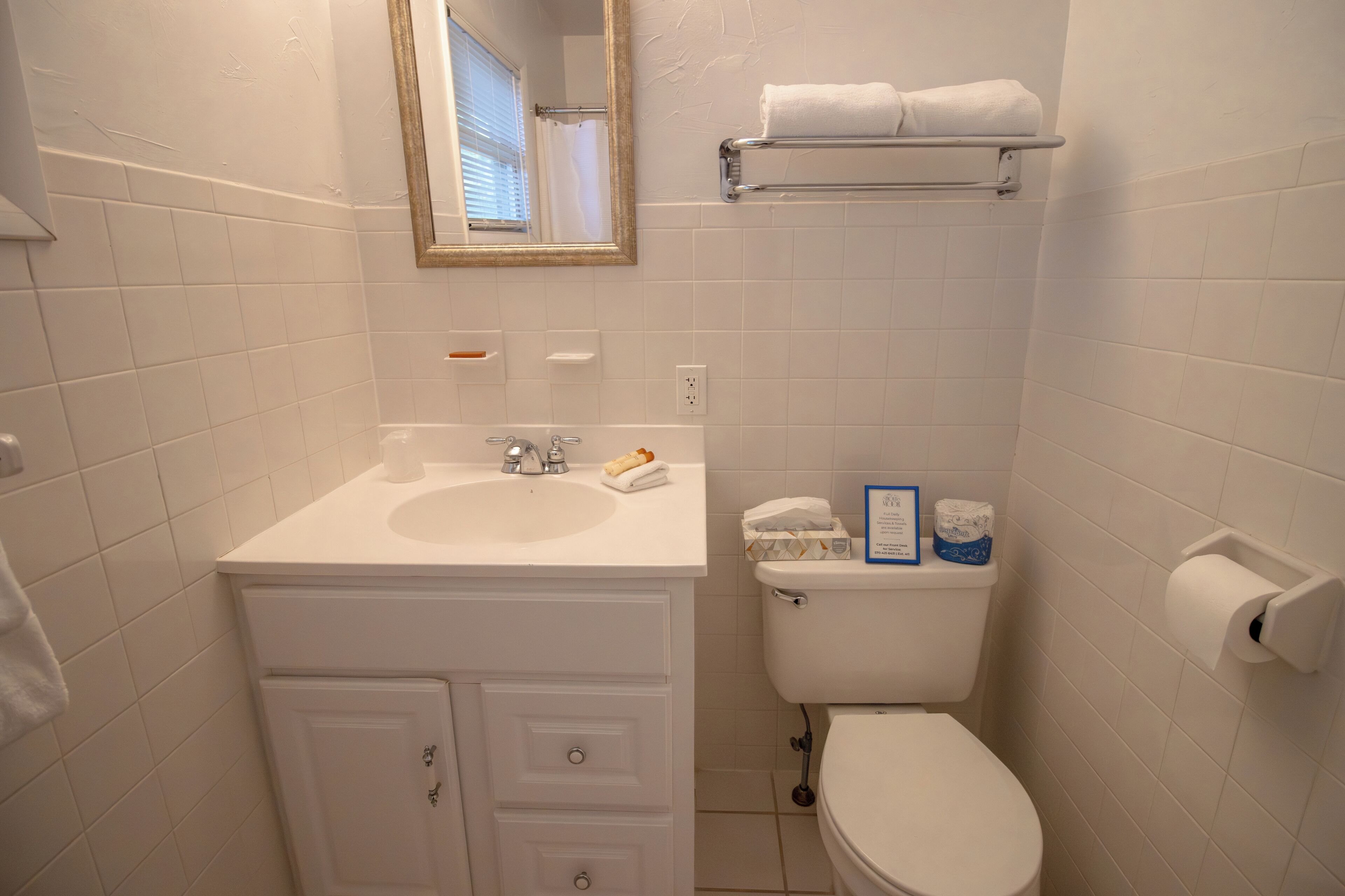 A bathroom with a white sink, mirror, and toilet. There is a towel rack above the toilet and a roll of toilet paper on the wall.