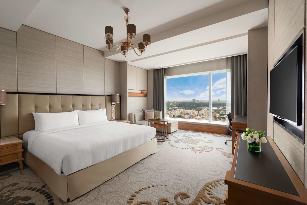 A modern hotel room with a large bed, a desk, a TV, and a view of the city.