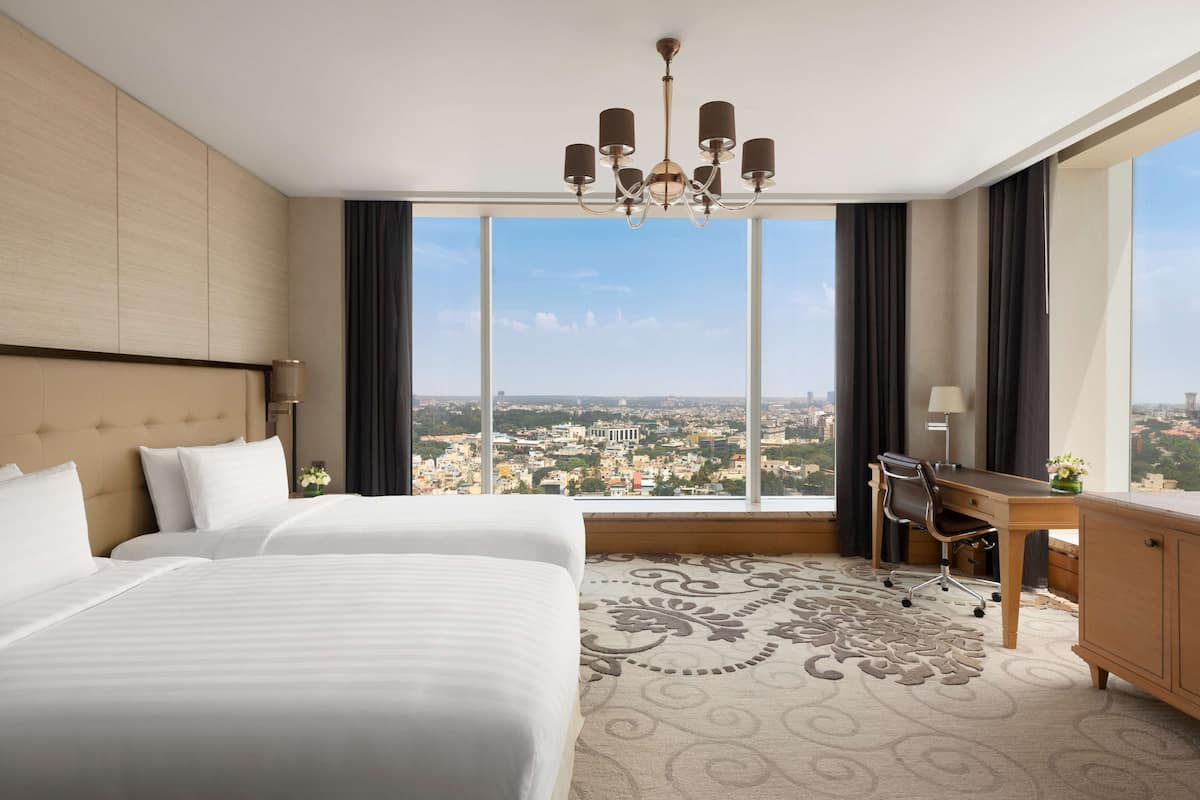 A hotel room with two beds, a desk, a chair, a chandelier, and a large window with a city view.