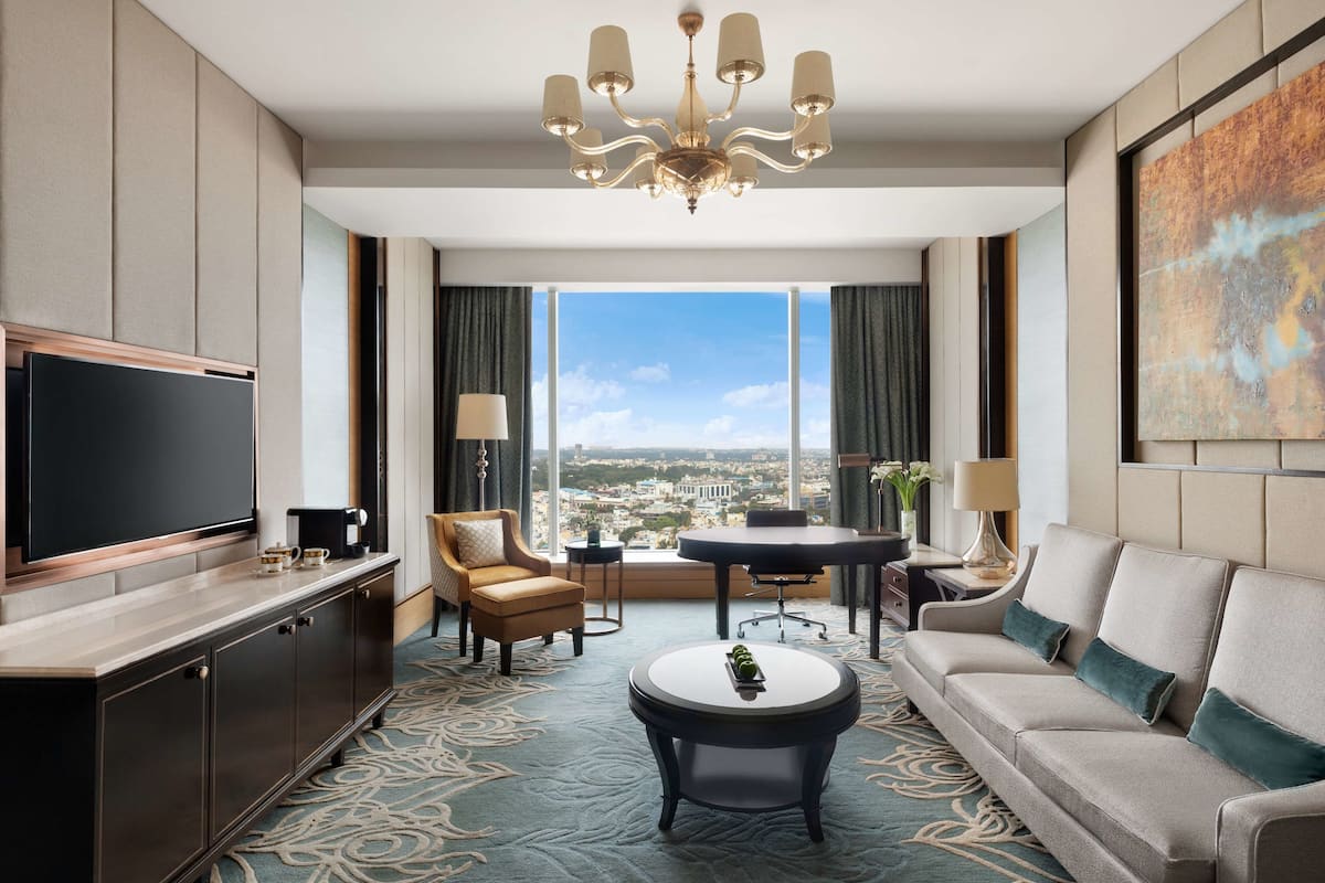 A modern hotel room with a large window, a flat-screen TV, a sofa, a coffee table, and a dining area.