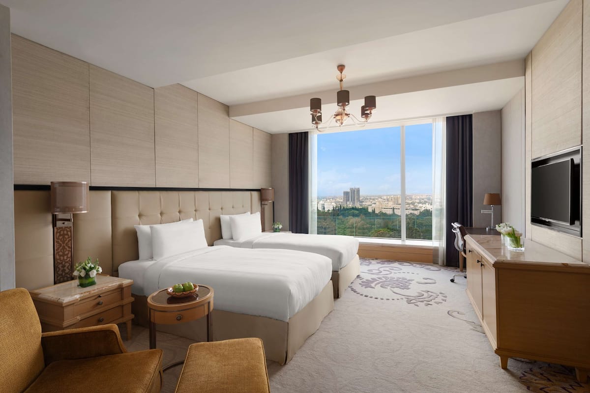 A modern hotel room with two beds, a TV, a desk, and a view of the city.