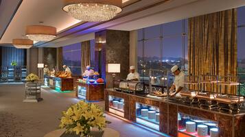 A modern hotel lobby with a bar area, chefs preparing food, and a view of the cityscape at night.
