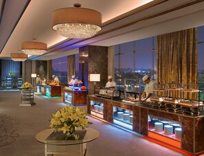 A modern hotel lobby with a bar area, chefs preparing food, and a view of the cityscape at night. - ARTEZIA (Bengaluru)