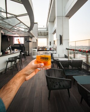 A person holding a glass with "Hype" on it, in a modern bar area with high ceilings and a city view. - Shangri-La Bengaluru (Bengaluru)