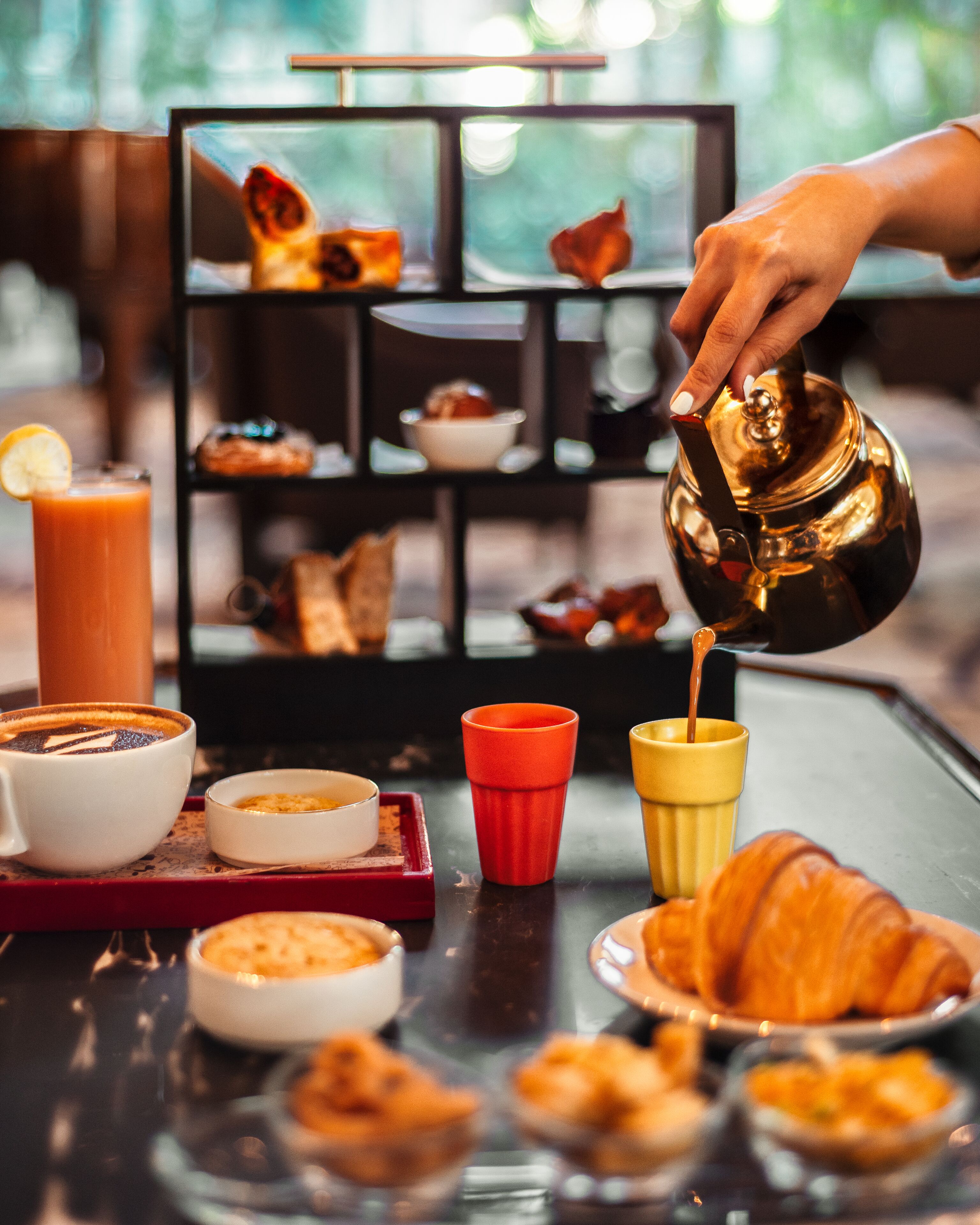 Une table mise pour le petit-déjeuner avec une tasse de café, un verre de jus d’orange, un croissant et une théière d’où s’écoule le thé.