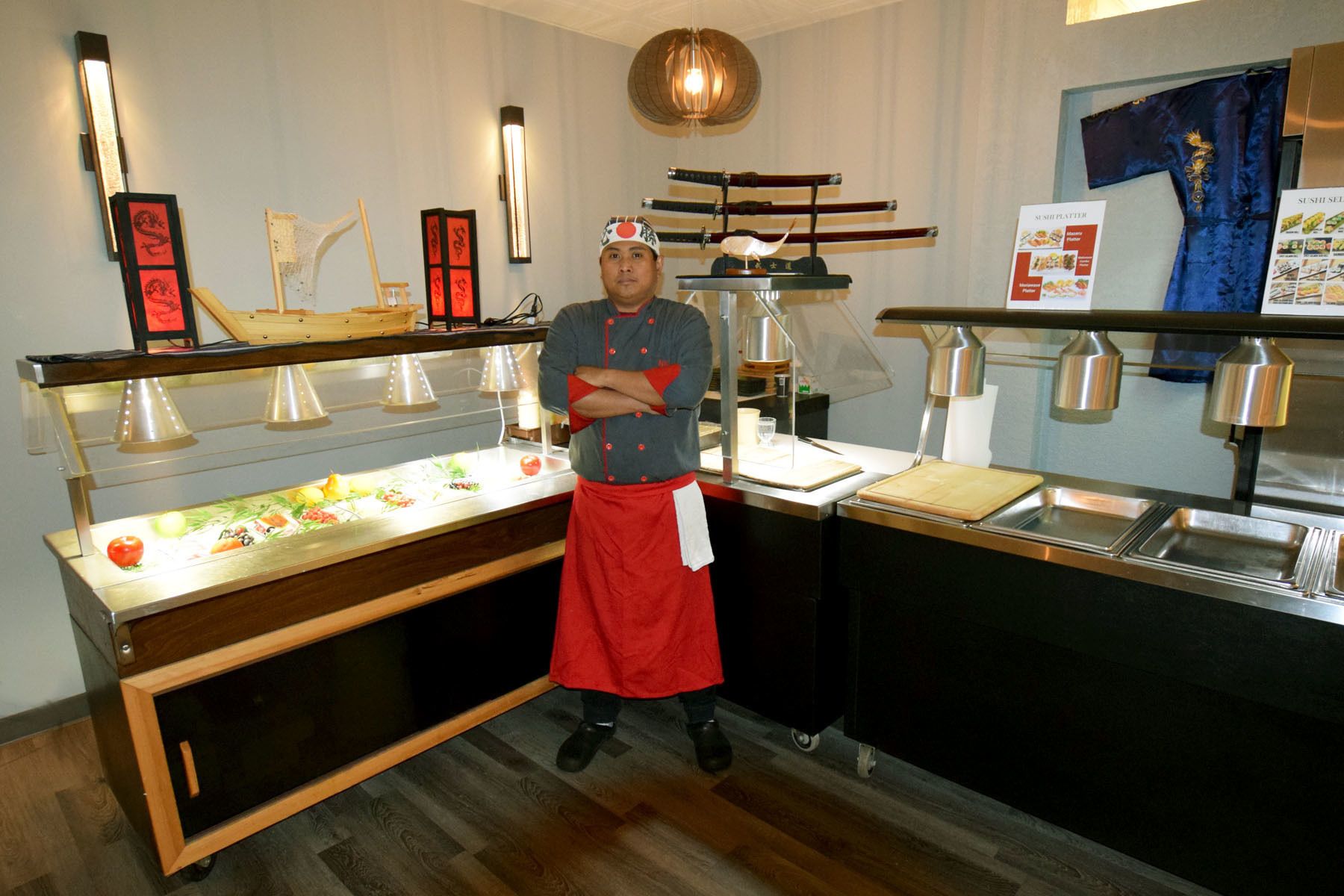 A chef standing in a kitchen with a sushi display and a traditional Japanese sword.