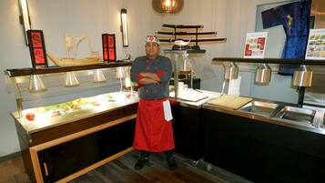A chef standing in a kitchen with a sushi display and a traditional Japanese sword.