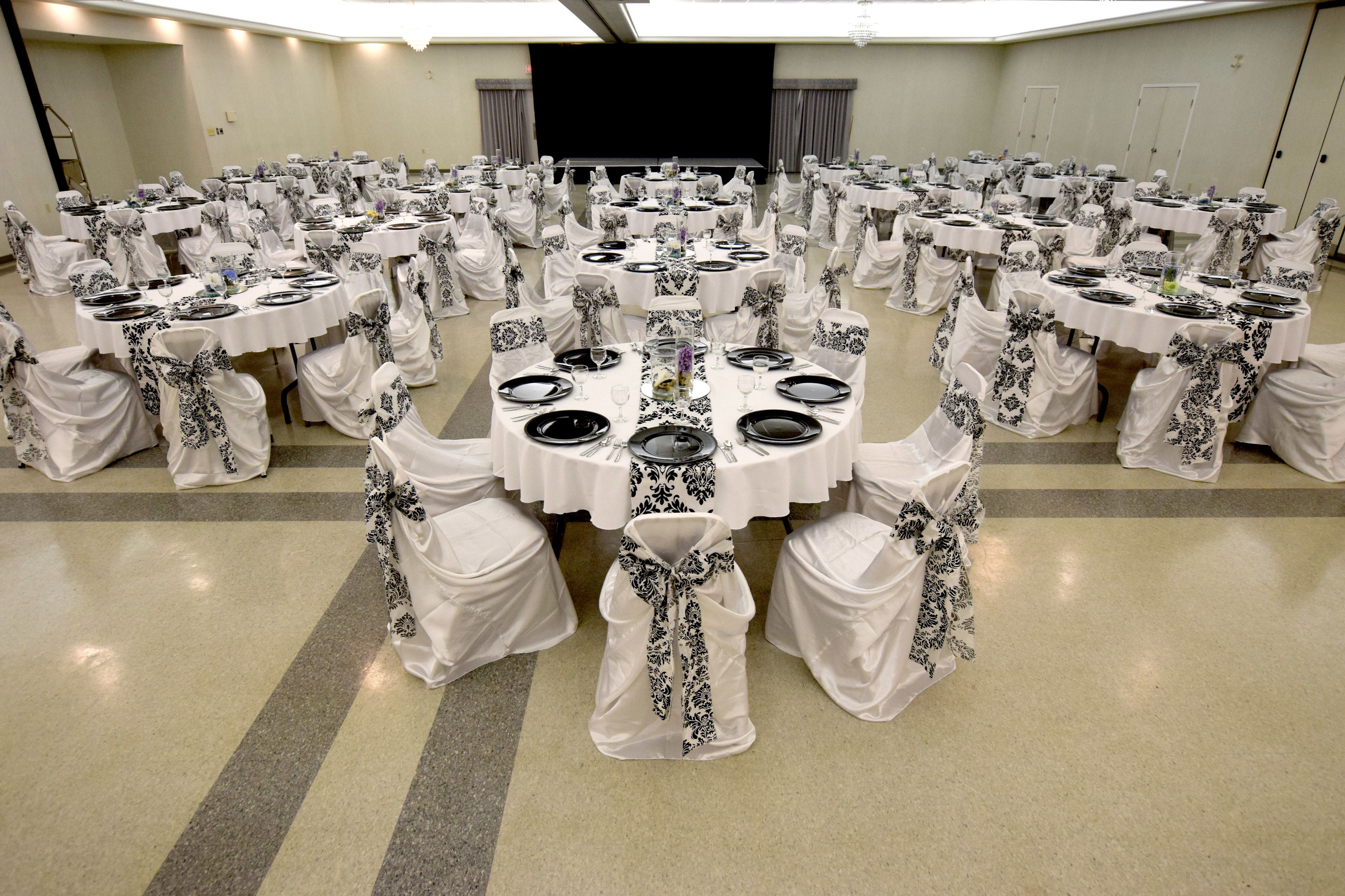 A banquet hall with round tables set for a formal event, featuring white tablecloths and black and white chair covers.