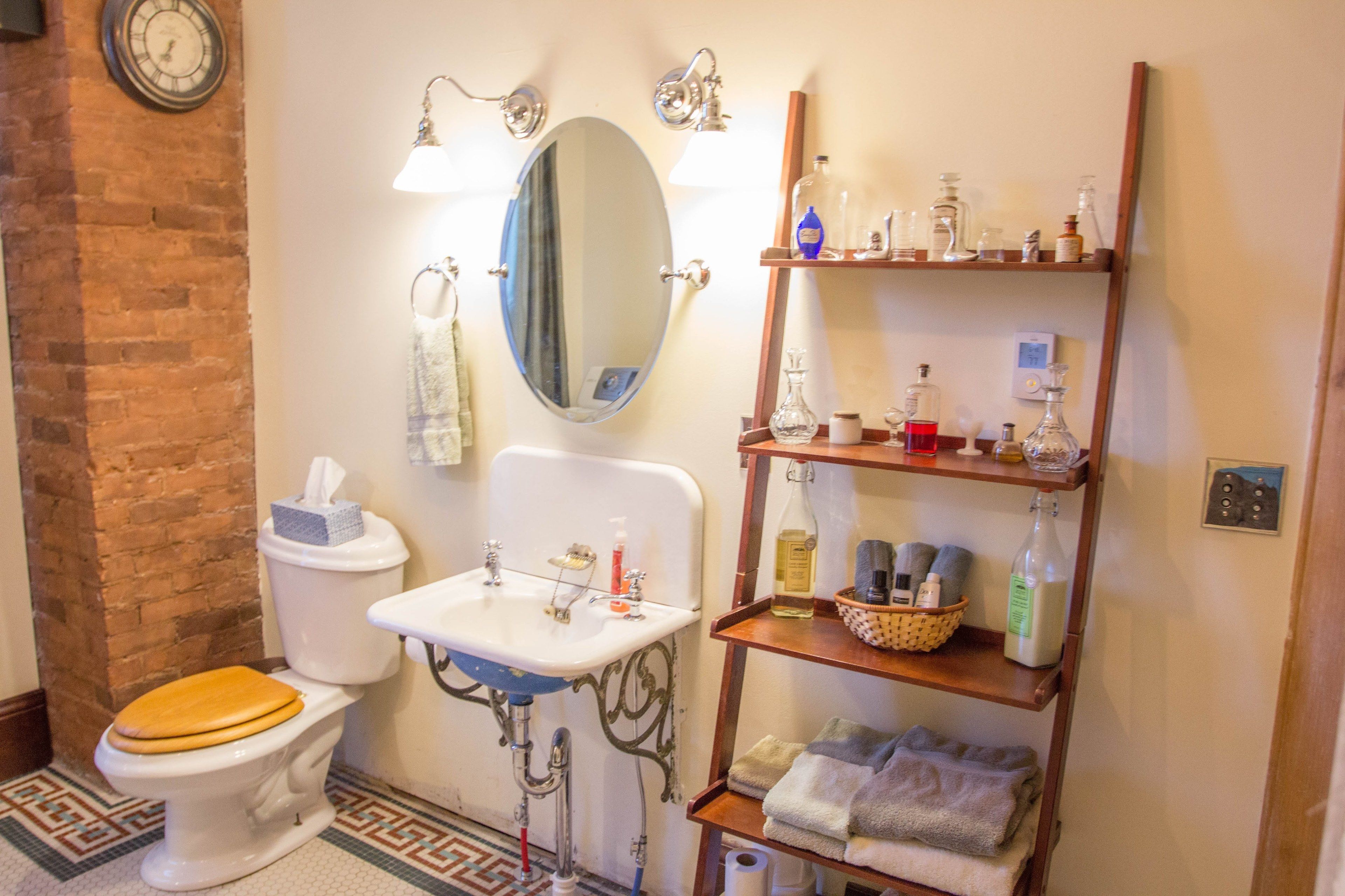 A bathroom with a toilet, sink, mirror, shelves with toiletries, and a clock on the wall.