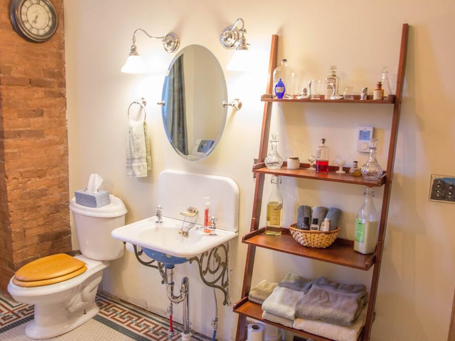 A bathroom with a toilet, sink, mirror, shelves with toiletries, and a clock on the wall.