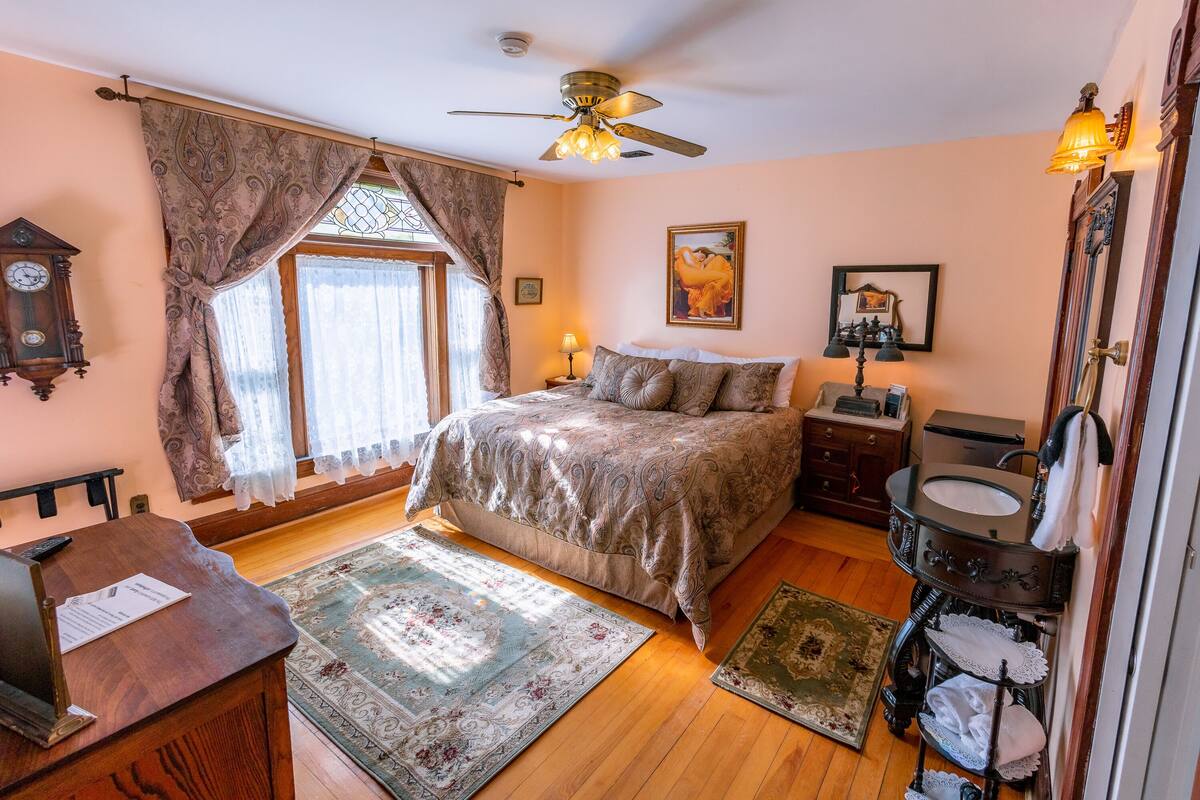 Une chambre à coucher comprenant un lit, un bureau, une commode, un miroir, une horloge et un ventilateur de plafond.