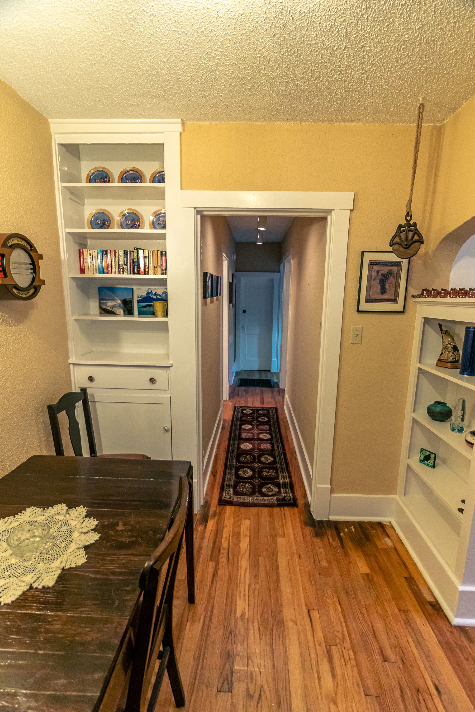 Un pasillo con suelo de madera, un comedor con mesa y sillas, y una estantería llena de libros y objetos decorativos.