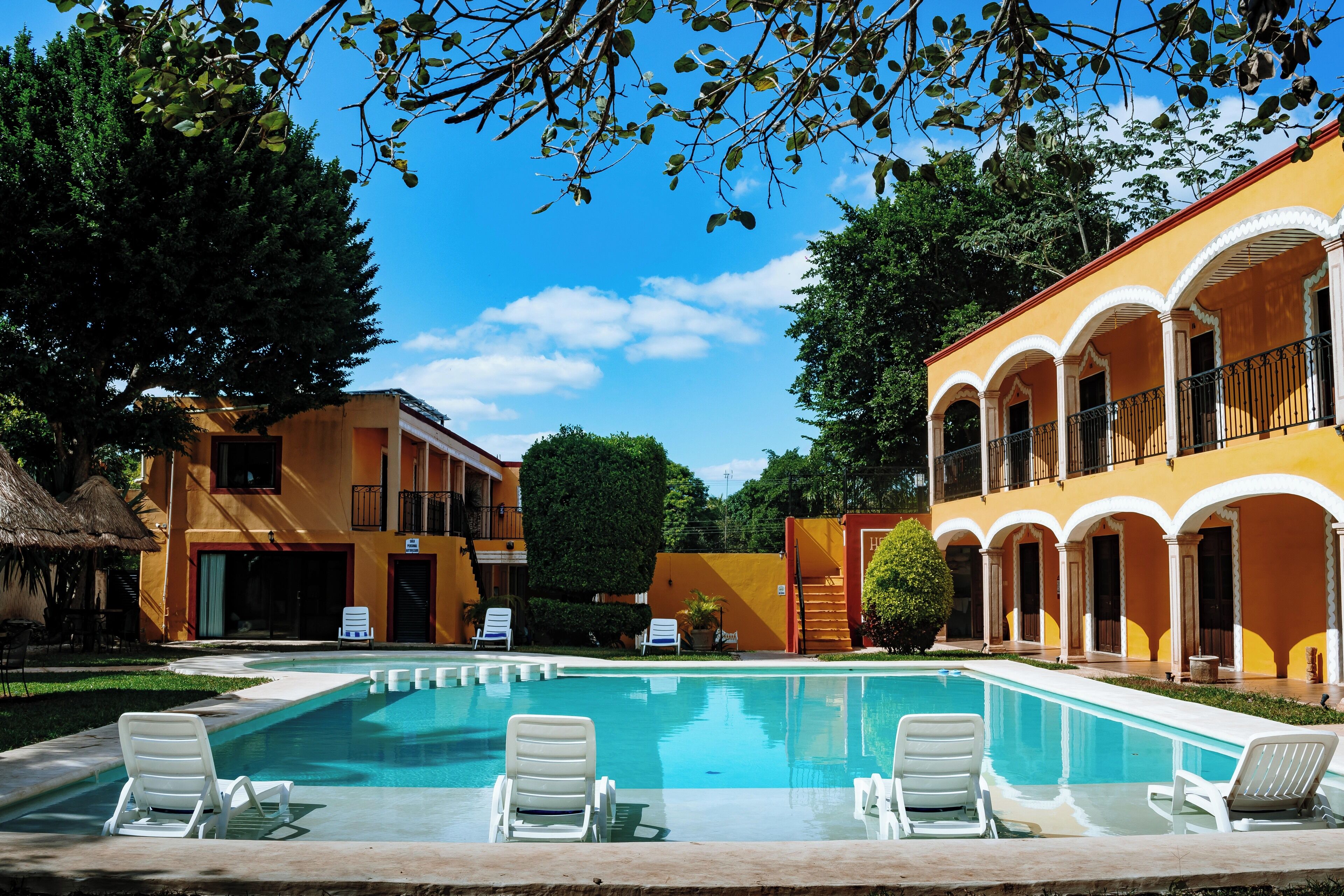A swimming pool with lounge chairs, a thatched-roof structure, and a building with yellow walls and arches.