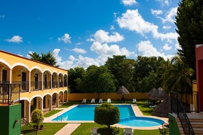 A pool area with a clear blue sky, a pool, lounge chairs, and a building with a yellow facade and white arches. - Hotel Tuul (Izamal)